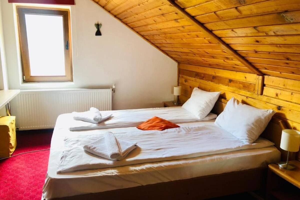 Un dormitorio con techo de madera inclinado, una ventana, un radiador, una cama con sábanas blancas y una mesita de noche con una lámpara.