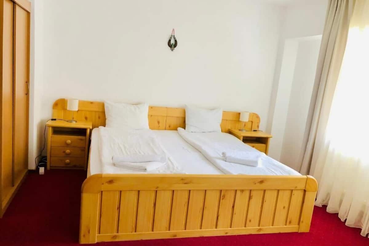 Una cama doble con cabecera y piecera de madera, dos mesitas de noche, una lámpara fijada a la pared y suelo cubierto de alfombra roja.
