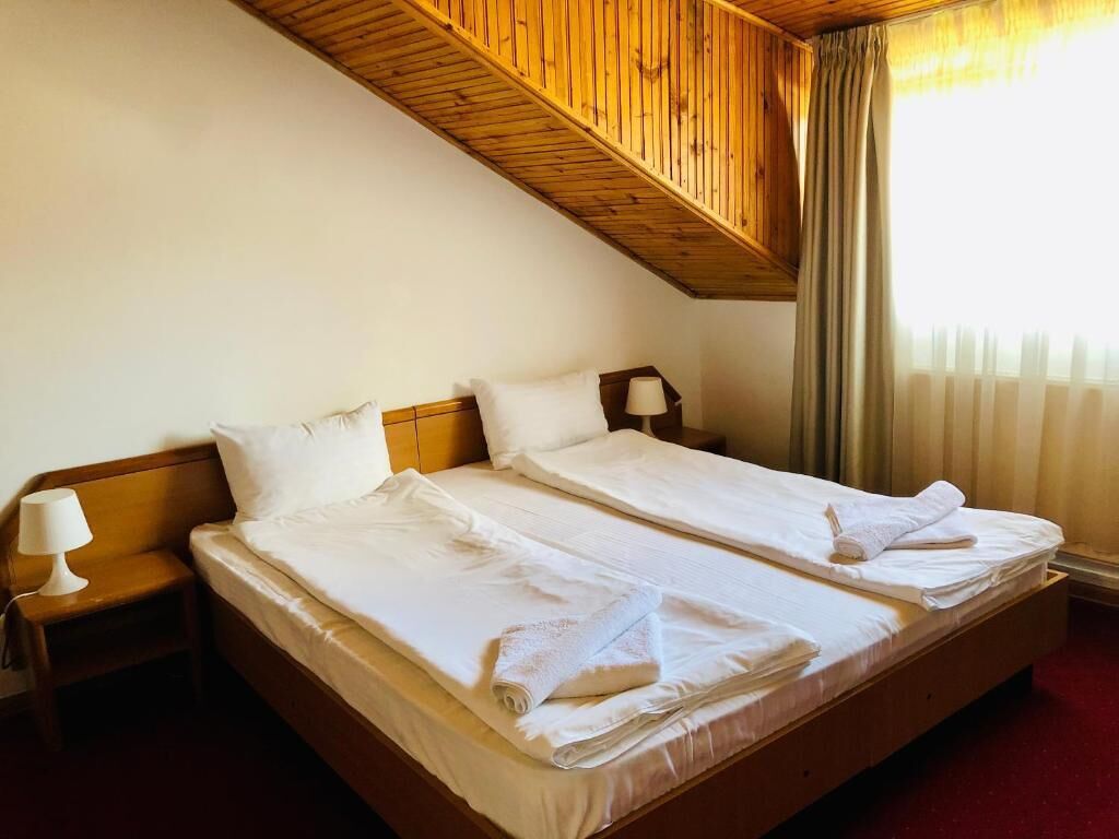 A hotel room with two beds, wooden headboards, a nightstand with a lamp, and a window with curtains.
