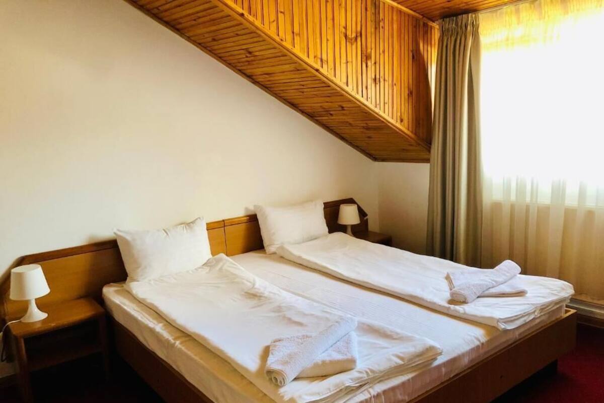 Habitación de hotel con dos camas, cabeceros de madera, una mesita de noche con lámpara y una ventana con cortinas.