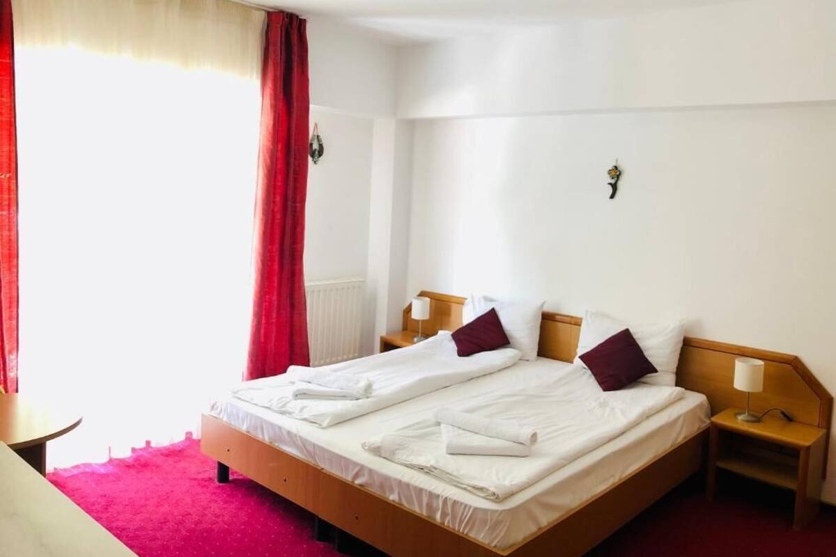 Una cama doble con dos almohadas, una mesita de noche con lámpara y una ventana con cortinas rojas.
