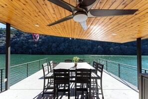 Un espacio exterior techado para comer con mesa y sillas, un ventilador de techo y vista a un lago.