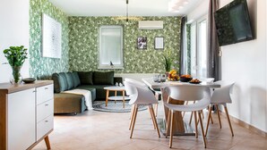 A modern living room with a green leaf-patterned wallpaper, a green sofa, a dining table with white chairs, and a kitchen area with a window and a radiator.