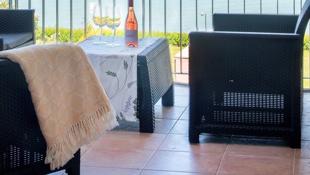A balcony with two wicker chairs, a table with a bottle and glasses, and a view of a body of water and distant hills.
