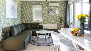 A modern living room with a green sofa, a round coffee table, and a dining area with white chairs and a fruit bowl.