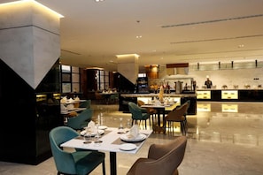 A modern dining area with tables set for service, a kitchen in the background, and a clean, polished floor. - Novo Hotel Riyadh (Riyadh)