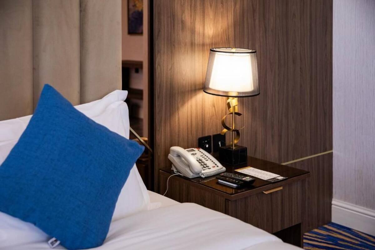 Una habitación de hotel con una cama, un almohadón azul, una mesita de noche con una lámpara, un teléfono y un control remoto.