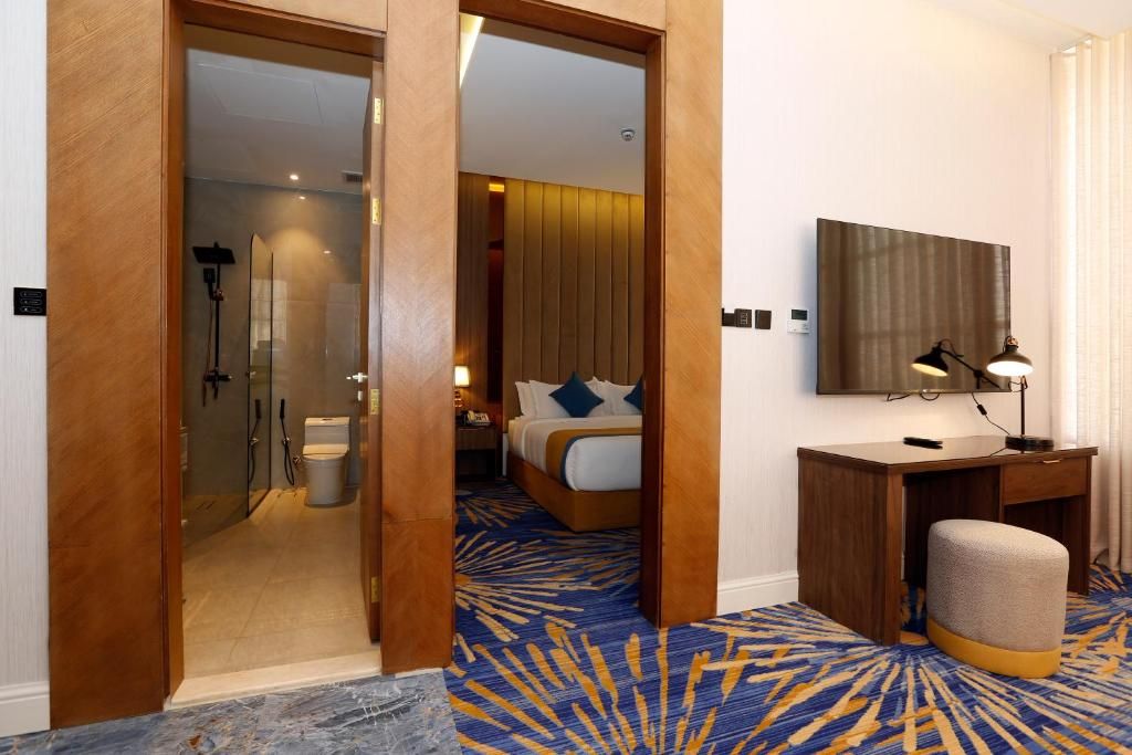 Habitación de hotel con puerta de vidrio que da a un baño, una cama con alfombra de diseño azul y amarillo, un escritorio con lámpara y un espejo grande en la pared.