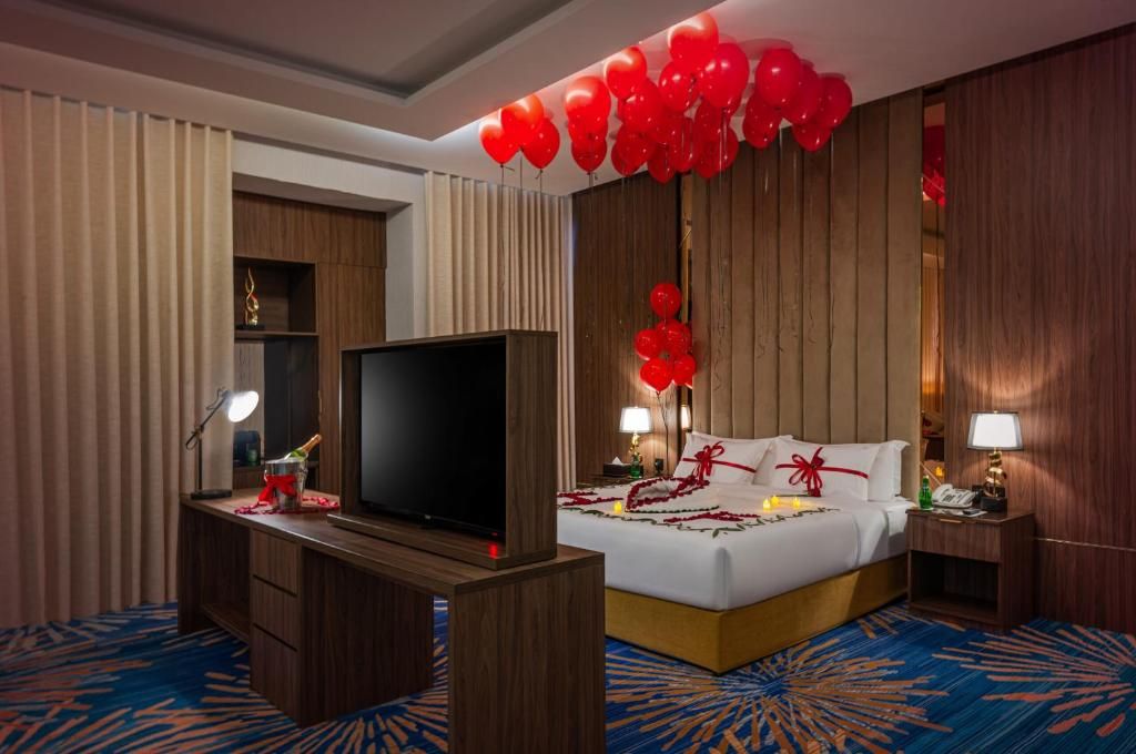 A hotel room with a bed, a flat-screen TV, a desk with a lamp, and a wall-mounted shelf.