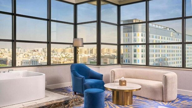 A modern hotel room with a large window offering a city view, a white bathtub, a blue armchair, a beige sofa, and a round coffee table. - Novo Hotel Riyadh (Riyadh)