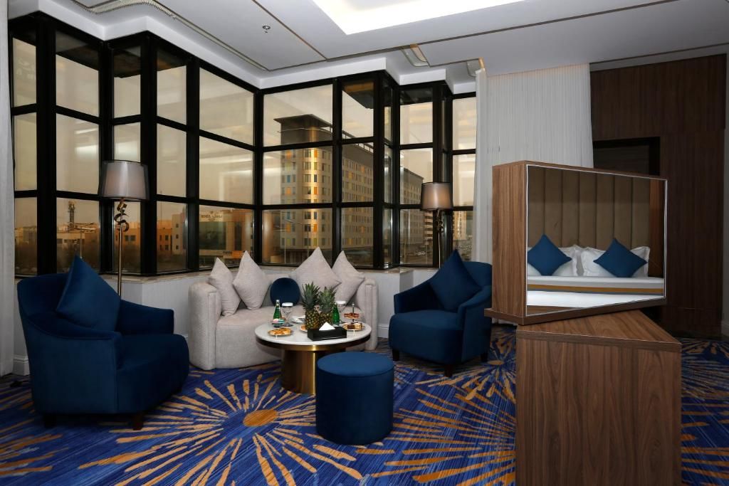 Habitación de hotel con un ventanal grande, un sofá, dos sillones, una mesa redonda de centro y un armario de madera con espejo.