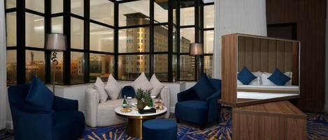 Habitación de hotel con un ventanal grande, un sofá, dos sillones, una mesa redonda de centro y un armario de madera con espejo.