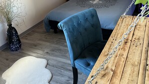 A bedroom with a bed, a blue chair, a wooden table, a wall-mounted shelf, and a decorative wall panel.