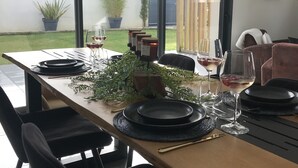 A dining area with a table set for two, featuring black plates, cutlery, and wine glasses. There is a clock on the wall and potted plants in the background.
