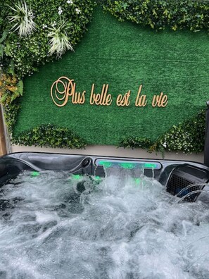 Boblebad med vandstråler, omgivet af frodig vegetation og en mur med teksten "Plus belle est la vie".