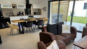 A modern kitchen with a dining area, a brown leather sofa with a deer pillow, and a view of a grassy yard through glass doors.