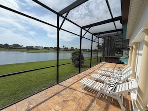 A covered outdoor area with a view of a lake, white lounge chairs, and a patio with stone tiles.
