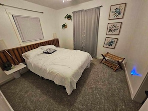 A bedroom with a bed, a bedside table, a bench, a window with blinds, and wall-mounted artwork.