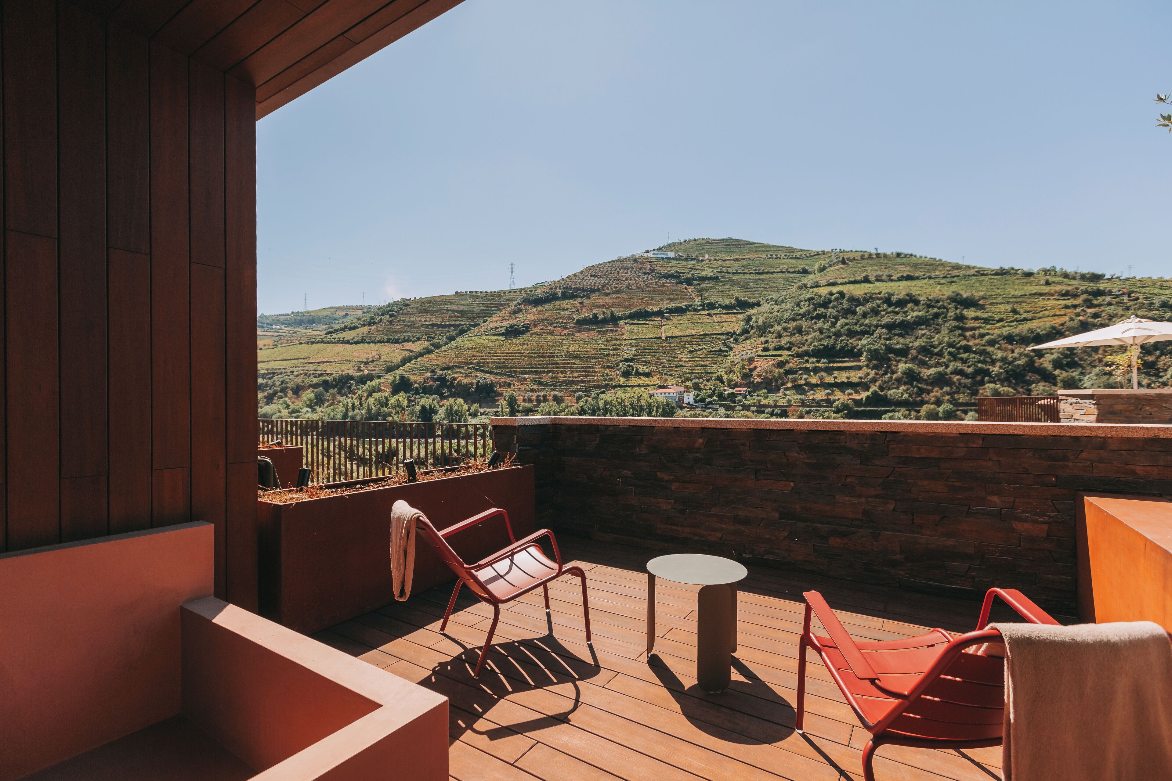 A wooden deck with red chairs and a small table, overlooking a hilly landscape.