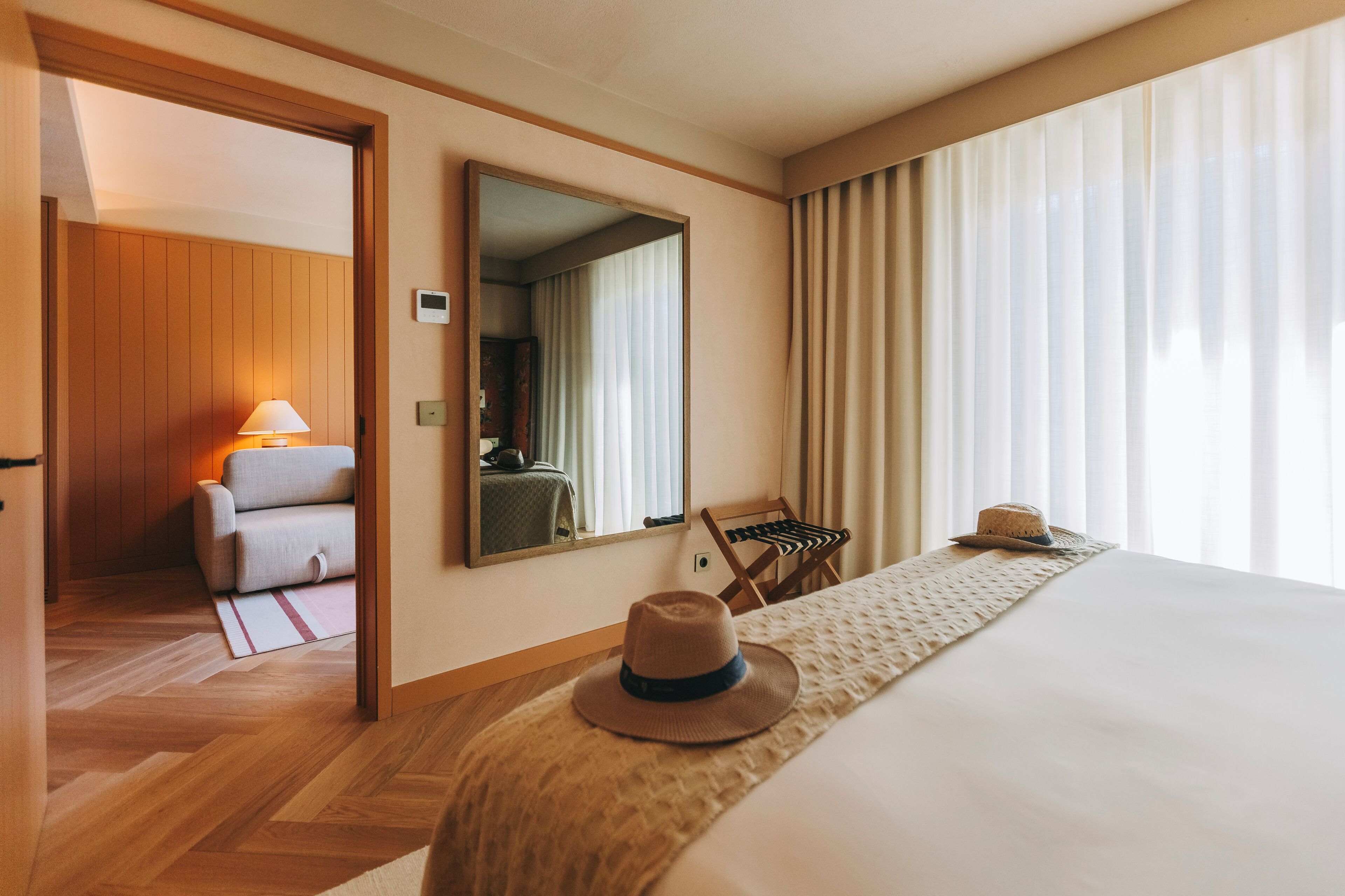 A hotel room with a bed, a mirror, a chair, and a hat on the bed.
