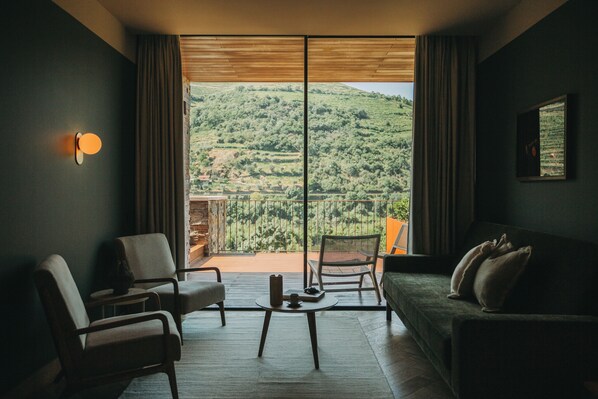 A modern living room with a sofa, two armchairs, a coffee table, and a view of a hilly landscape through large windows.