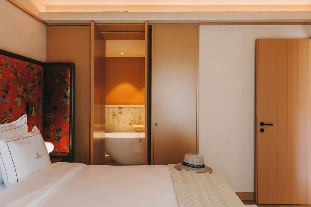 Habitación de hotel con cama, mesita de noche, un sombrero, una puerta de madera y un baño con bañera.