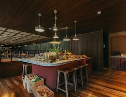 A restaurant with a wooden ceiling, hanging pendant lights, a central island with food and drinks, and wooden stools.