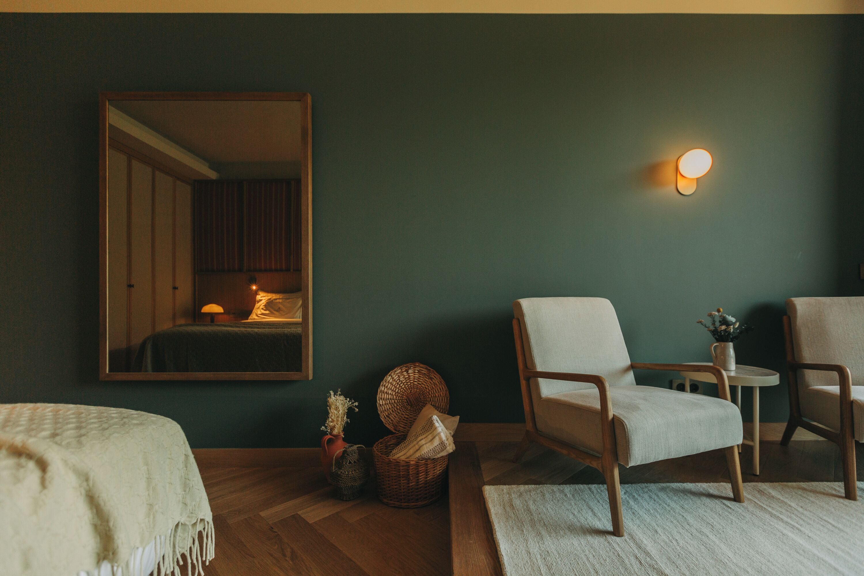 A room with a bed, two armchairs, a small table, a mirror, and a wall-mounted light fixture.