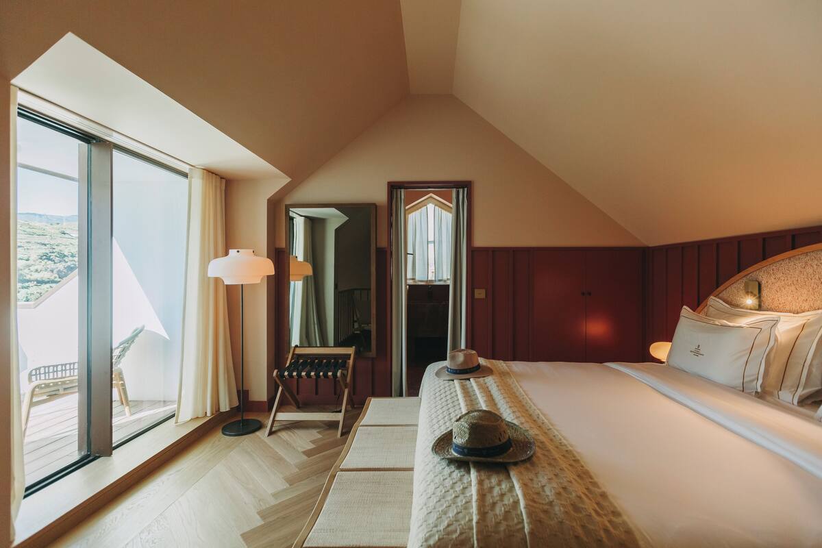 Habitación de hotel con un ventanal grande, una cama con almohadas, un sombrero sobre la cama, una silla y una lámpara.