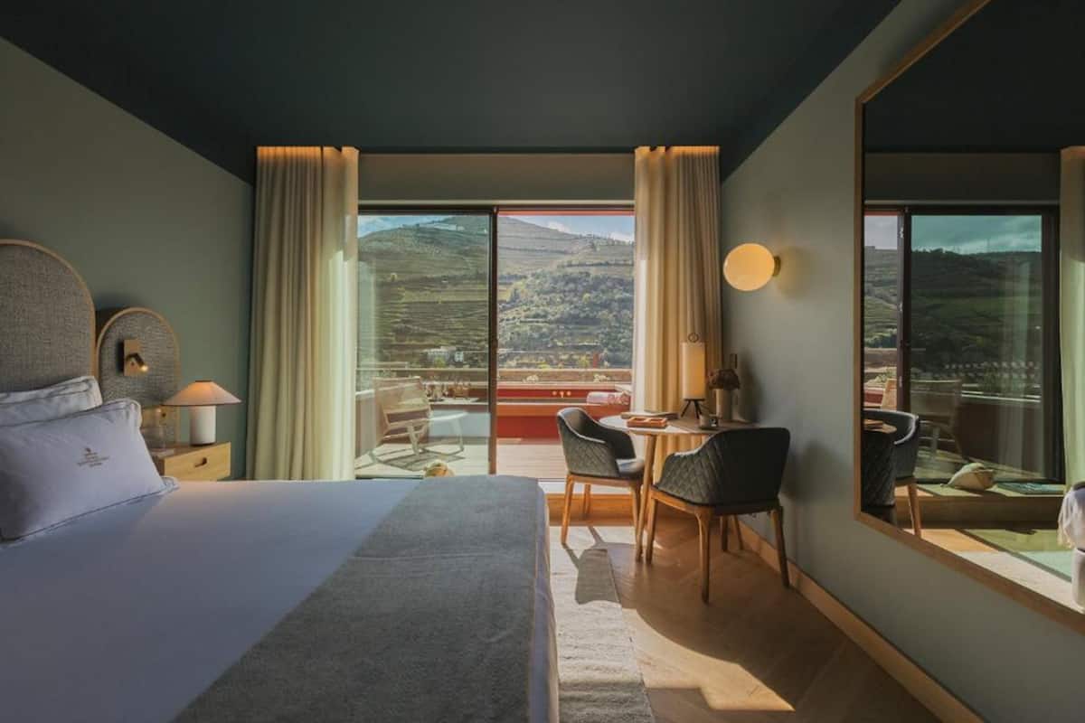 Habitación de hotel con una cama grande, un escritorio, sillas y vistas al campo a través de amplias ventanas.