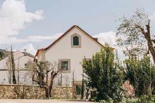 Un edificio blanco con techo de tejas rojas, una ventana pequeña y un muro de piedra al frente.
