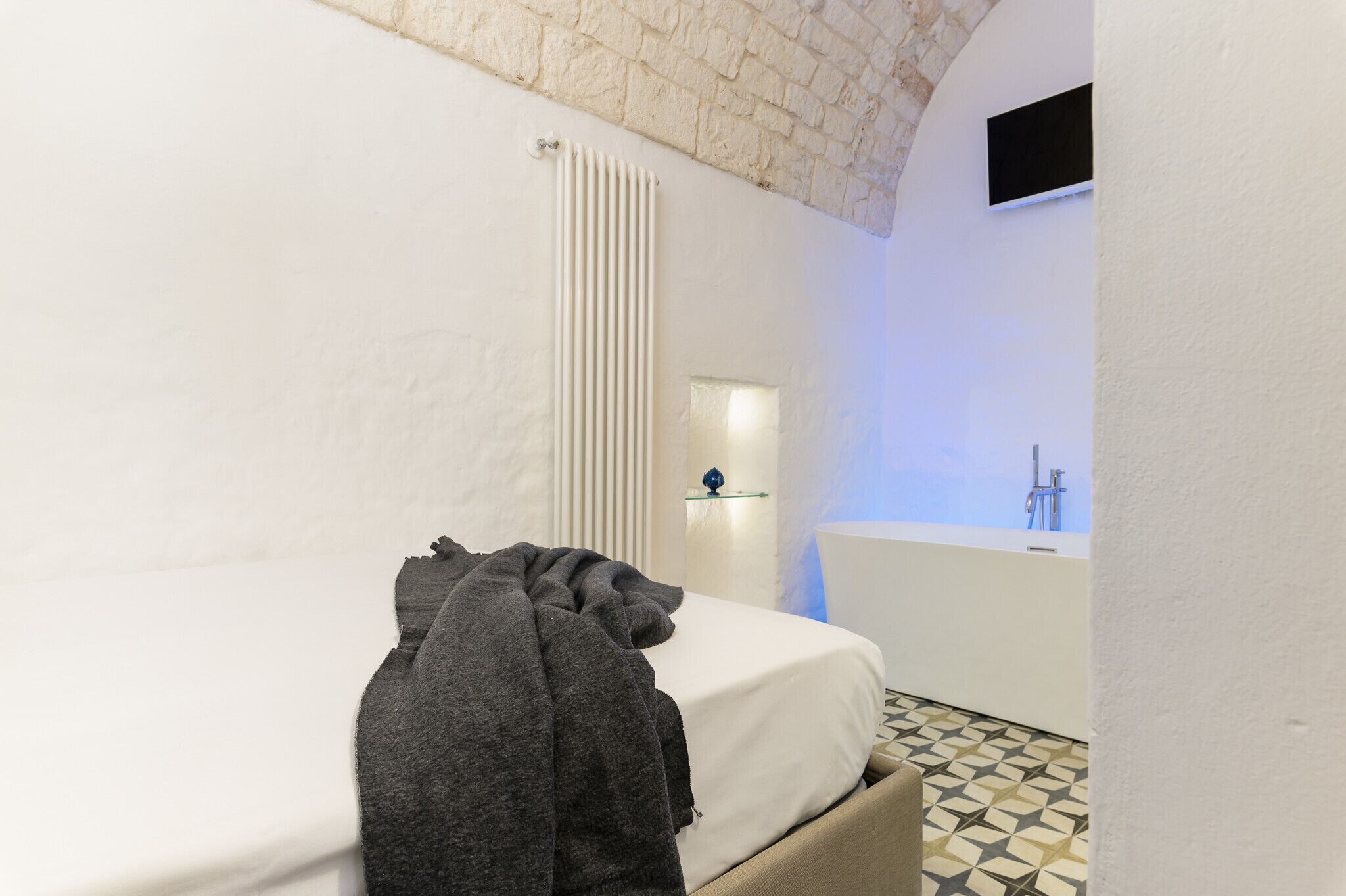 A bedroom with a bed, a radiator, a small table with a blue object, and a bathroom with a sink and mirror.