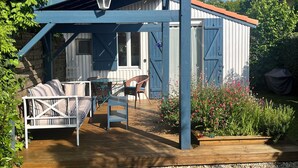 A covered outdoor seating area with a wooden deck, a white sofa, and a table. - Cabane Edena, Terrasse Ombragée Jardin Vélos Wifi à 350m de la Plage du Mauret (Andernos-les-Bains)