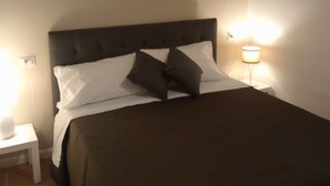Una cama bien hecha con cabecera oscura, almohadas blancas y colcha marrón. Hay una mesita de noche pequeña con una lámpara y un cuadro colgado en la pared sobre la cama.