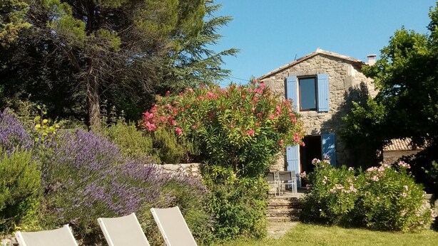 A stone house with a blue shutter, surrounded by lush greenery and flowering plants. - Charme D'un mas en Luberon, Calme, vue Panoramique, Piscine, Cheminée (Lacoste)