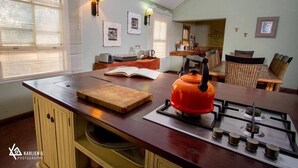 A cozy kitchen with a gas stove, a cutting board, and a kettle on the counter. There are wicker chairs and a dining table in the background. - Thorn Tree House (Livingstone)