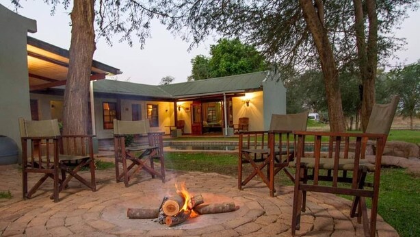 A patio area with a fire pit surrounded by wooden chairs, a building with a covered porch, and a well-maintained lawn. - Thorn Tree House (Livingstone)