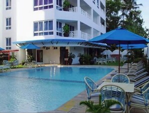 A swimming pool with lounge chairs and umbrellas, surrounded by a multi-story building with balconies.