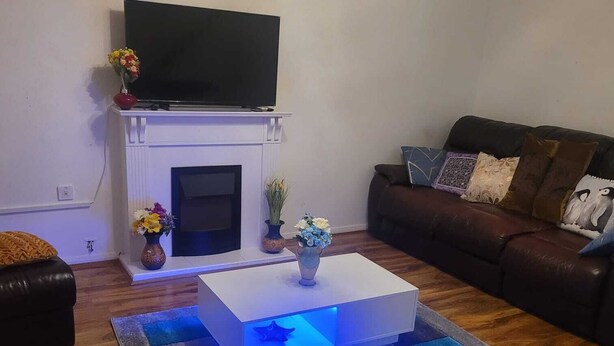 Un salon doté d’une cheminée, d’un téléviseur à écran plat fixé au-dessus, d’une table basse blanche avec un éclairage LED bleu, et d’un canapé en cuir marron agrémenté de coussins décoratifs.