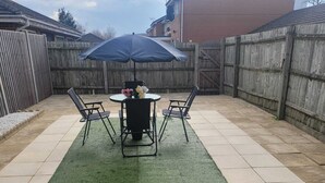 Une terrasse avec une table ronde et quatre chaises, une clôture en bois et un tapis de gazon synthétique vert.