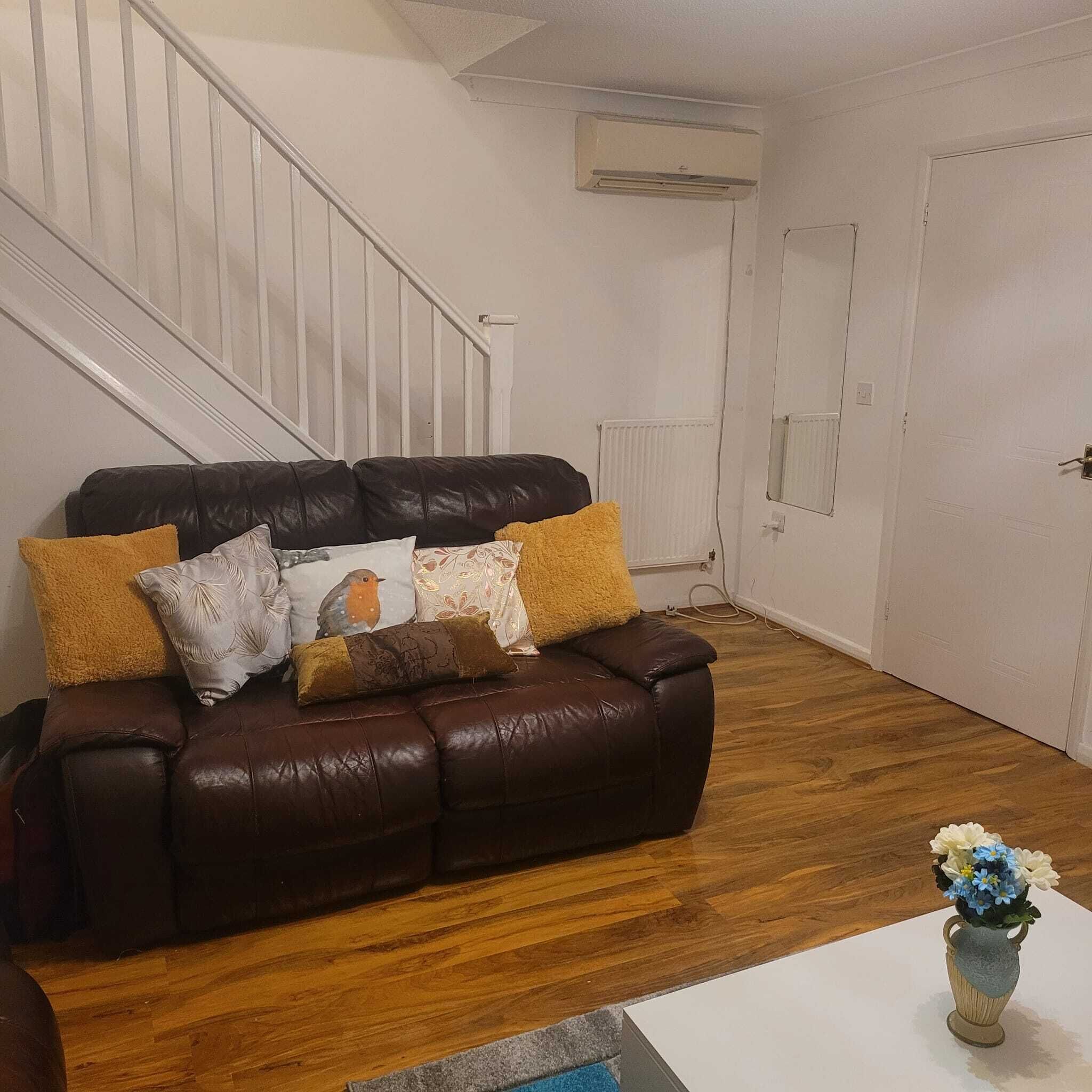 Un salon avec un canapé en cuir marron, un plancher en bois, une porte blanche et un petit vase avec des fleurs posé sur une table blanche.