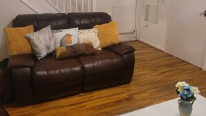 Un salon avec un canapé en cuir marron, un plancher en bois, une porte blanche et un petit vase avec des fleurs posé sur une table blanche.