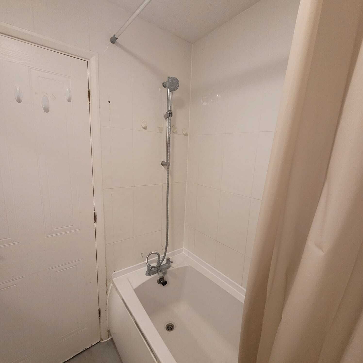 A small, white-tiled bathroom with a shower, a bathtub, and a door with three handles.
