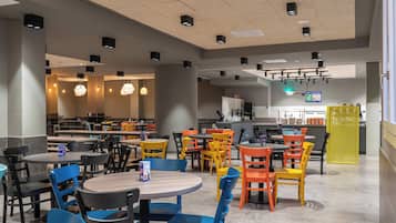 A spacious dining area with multiple tables and chairs, a kitchen area in the background, and a bright ceiling with recessed lighting.