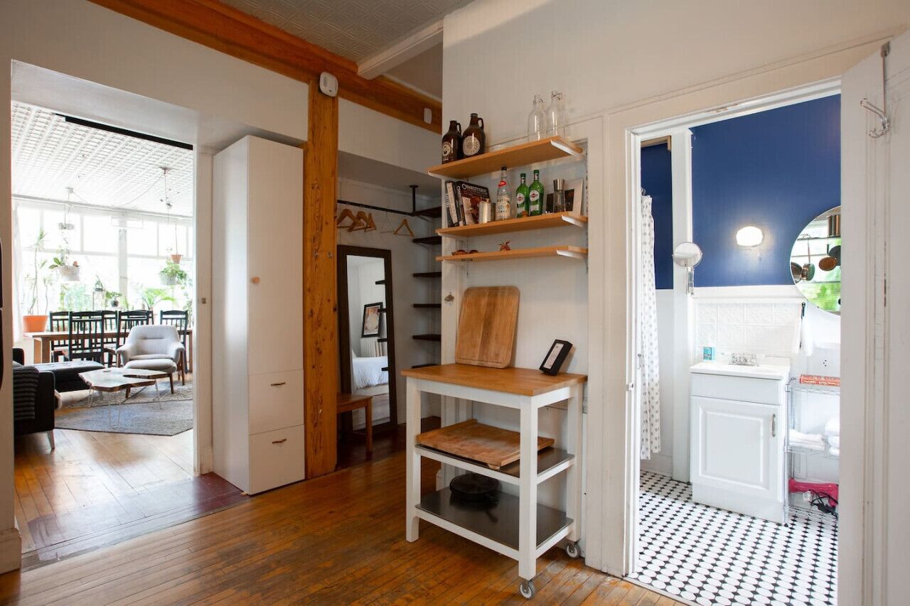 A room with a wooden floor, white walls, and a kitchen area with white cabinets and shelves.