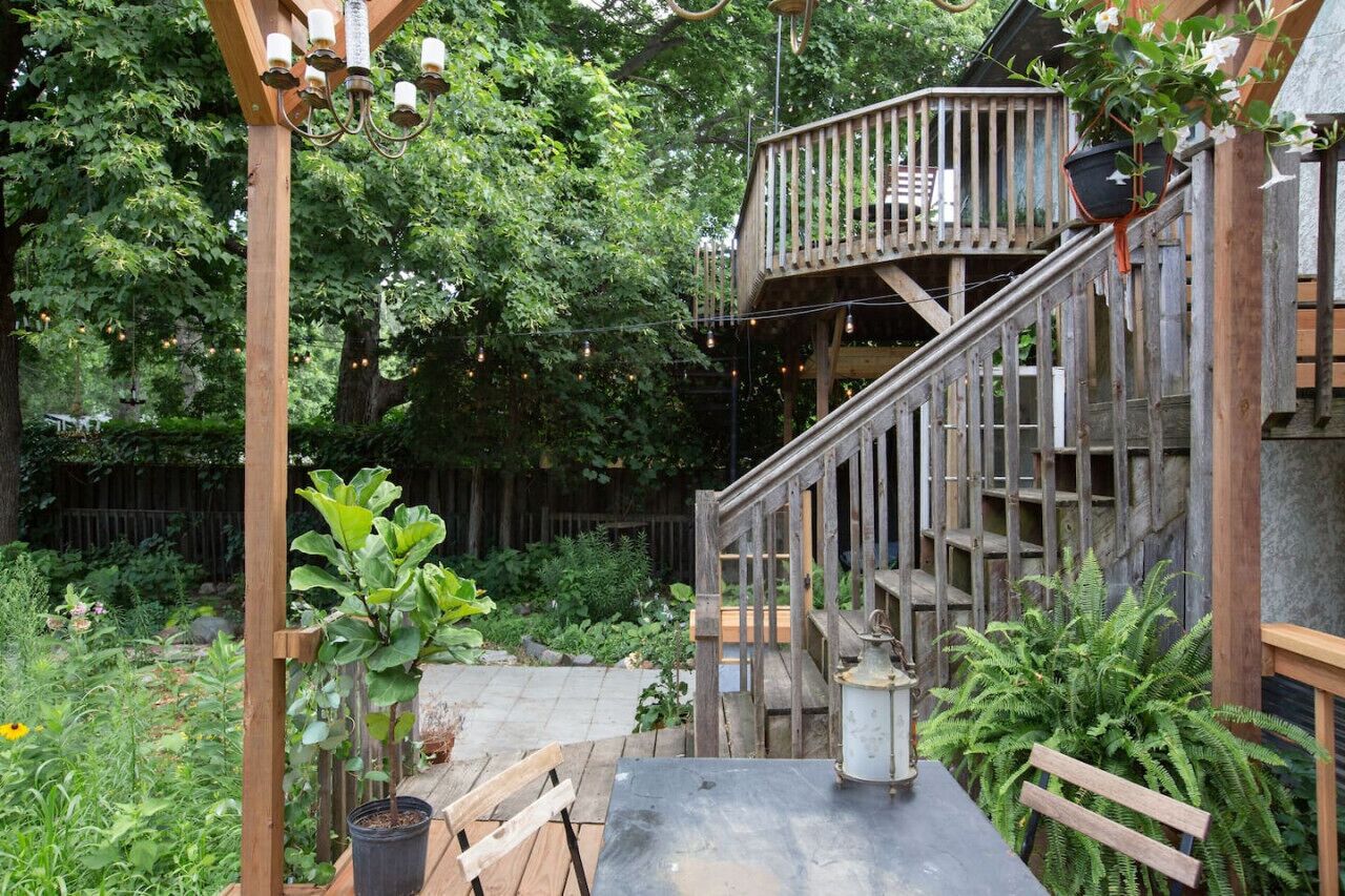 Una terraza de madera con una escalera que lleva a una plataforma elevada, rodeada de exuberante vegetación y un jardín.