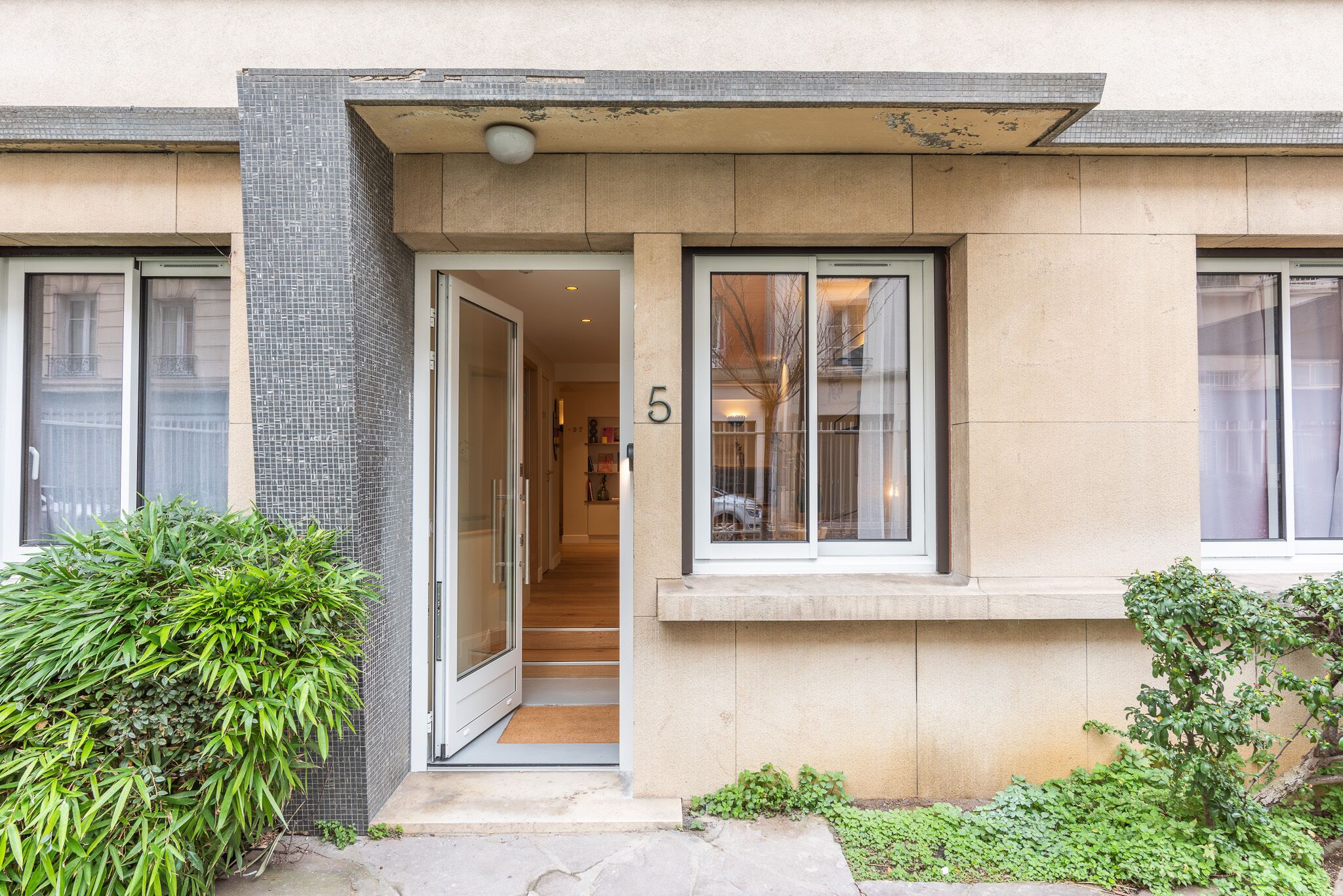 Un bâtiment moderne avec une façade recouverte de carreaux, un numéro « 5 » bien visible sur la porte et un petit hall d’entrée avec une marche.
