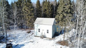 A small, single-story house with a sloped roof, surrounded by a snowy landscape and dense forest.