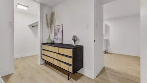 Ein schlicht eingerichteter Raum mit Holzboden, einem schwarzen und beigefarbenen Schminktischchen, einer Vase mit Pampasgras und einem gerahmten Bild an der Wand.