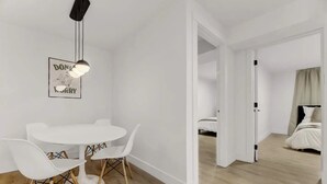 Ein moderner, minimalistischer Raum mit einem weißen Esstisch und Stühlen, einer Pendelleuchte und einem gerahmten Poster an der Wand.